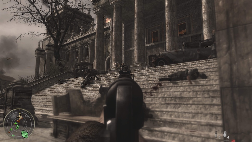 The player is aiming their MP40 sub-machine gun at German soldiers on the steps leading up to the Reichstag. On the right side there is a dead German soldier face down. On the left side there are a couple of more dead German soldiers and another that has just been shot in the chest and his holding his hand on the wound.